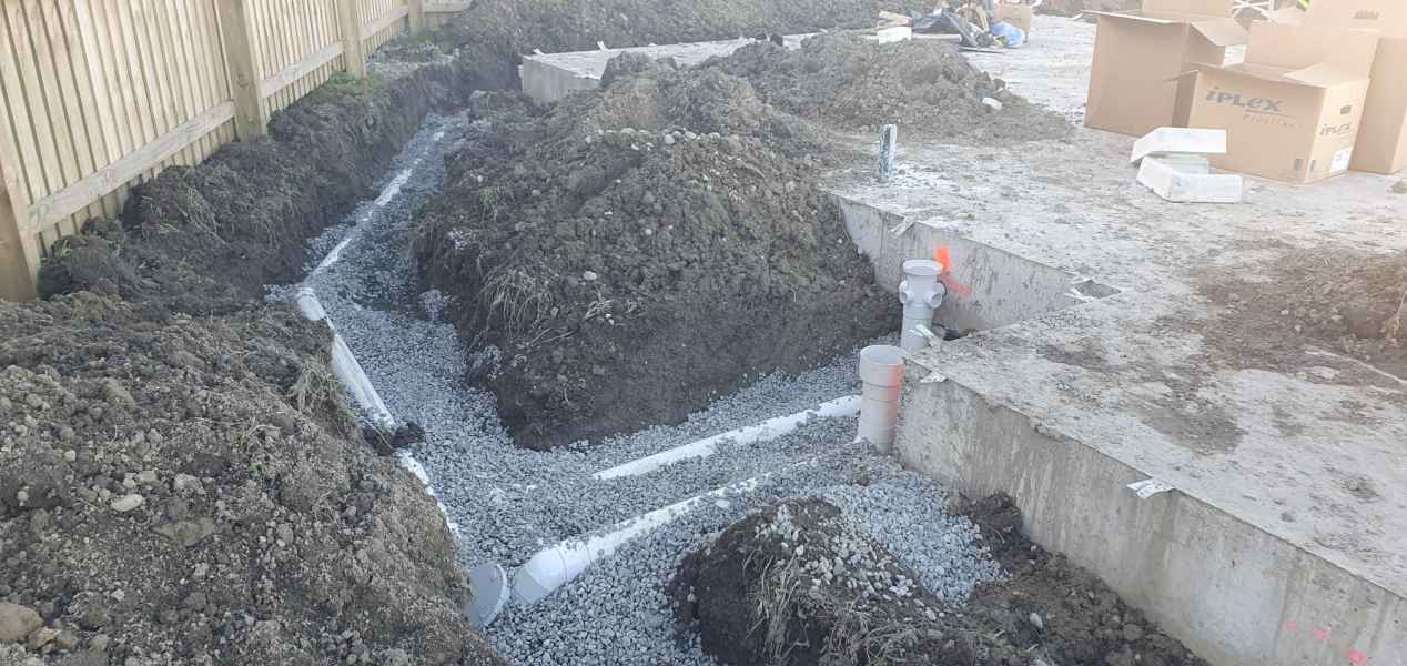 Gravel-filled trench with pipes installed for drainage at a construction site, surrounded by dirt and building materials.