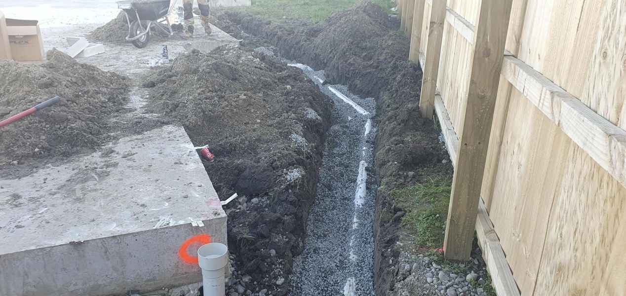 Excavated trench with gravel base for construction, featuring PVC pipe and surrounding soil on a building site.