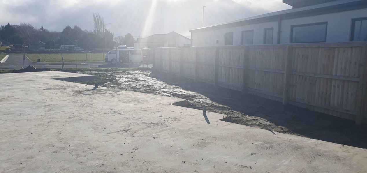 Construction site with freshly poured concrete floor and a trench for drainage, surrounded by a wooden fence and a clear sky