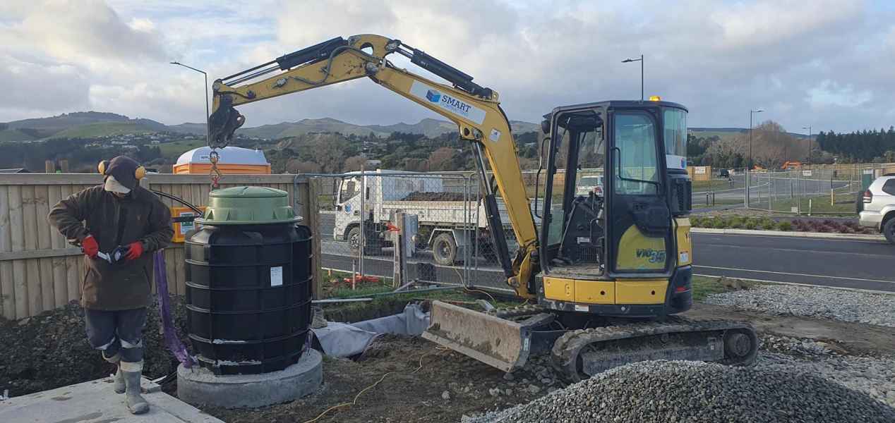 Excavator placing large drainage tank while a worker adjusts equipment at the construction site, with gravel piles and a fenced area in the background.