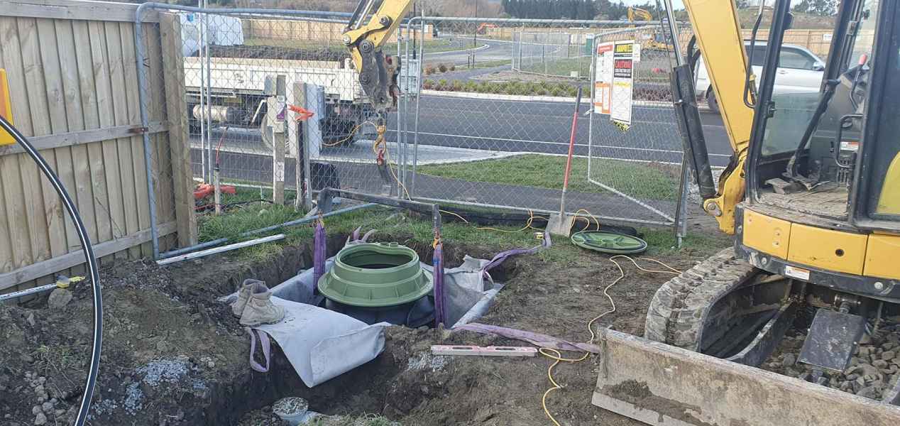 Excavator installing drainage system at construction site with green components in trench, supported by lifting straps