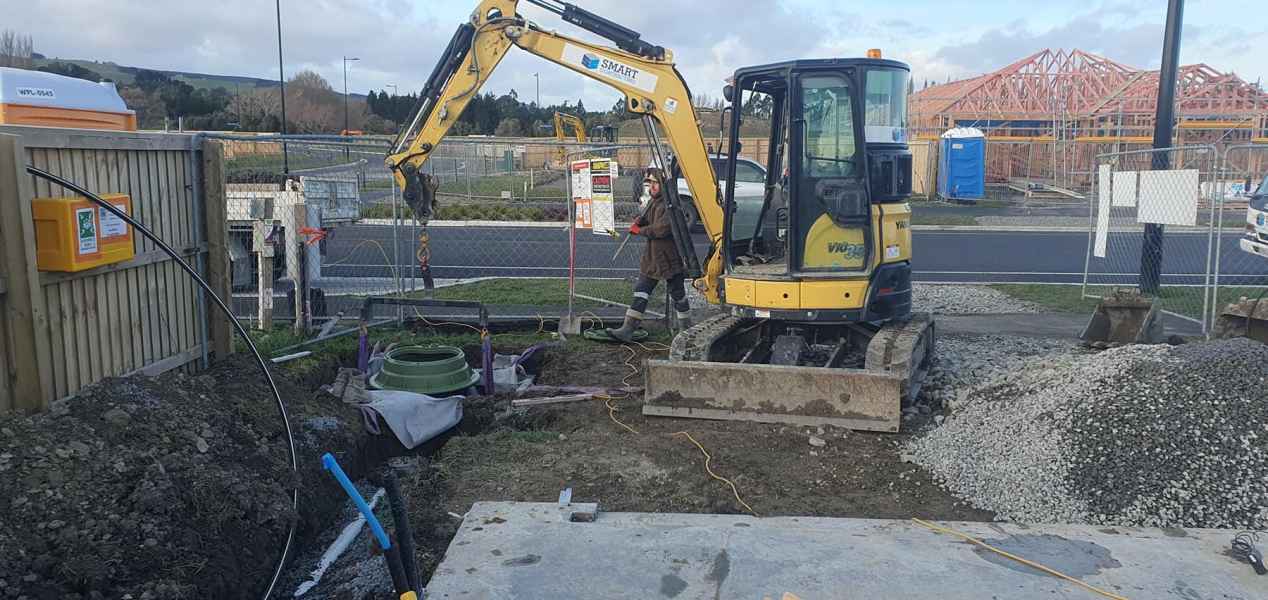 Excavator lifting green drainage component at a construction site with gravel piles and a worker walking nearby.