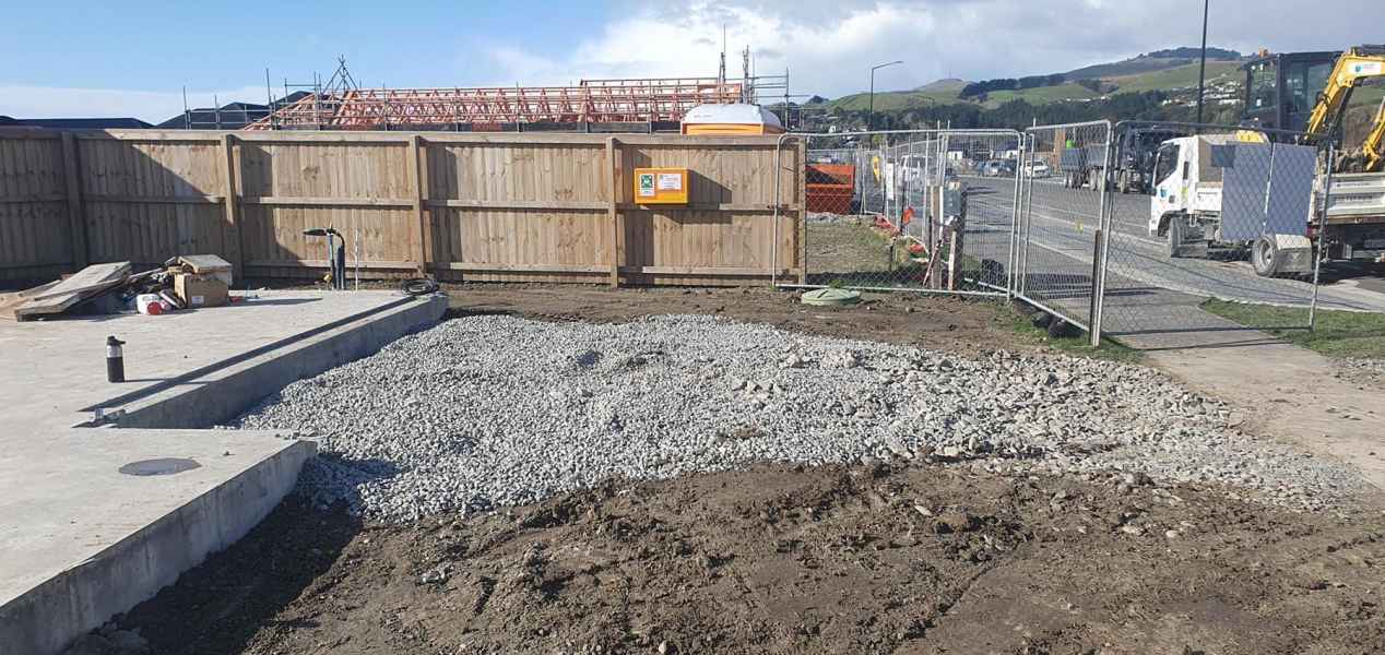 Construction site with gravel laid out on the ground, fenced area, and nearby construction materials, including a concrete slab
